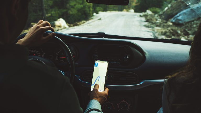 Fahrt in den Urlaub, Gadgets für lange Reisen im Auto Eine Person von hinten auf dem Fahrersitz eines Autos. Sie fährt auf einer Straße, während ein Handy an der Konsole die Route anzeigt.