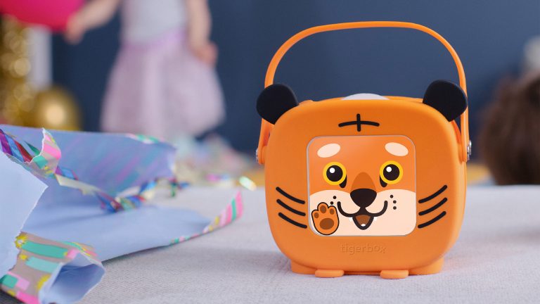 Tigerbox Mini, Süße Musikboxen kommen im September Eine Tigerbox Mini mit Tiger-Hülle steht auf einem Tisch neben Geschenkpapier und zeigt eine Winke-Animation an.
