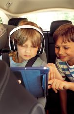 Zwei Kinder auf der Rückbank eines Autos schauen zusammen auf ein Tablet. Ein Kind trägt Kopfhörer.