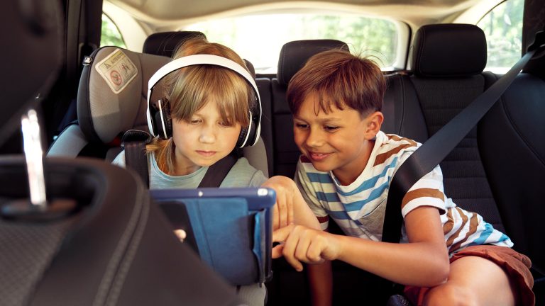 Reisen mit Kindern, Die besten Tipps für lange Autofahrten Zwei Kinder auf der Rückbank eines Autos schauen zusammen auf ein Tablet. Ein Kind trägt Kopfhörer.
