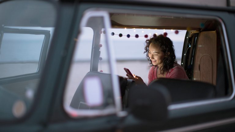 Camping Apps, Must-Haves für den Urlaub mit Camper oder Wohnmobil Blick durch das offene Fenster eines Campers, in dem eine Person an der offenen Seitentür sitzt und auf ihr Handy schaut.