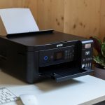 Der Epson EcoTank ET-2850 steht auf einem Schreibtisch. Im Hintergrund ist eine Holzwand und eine Pflanze zu sehen.