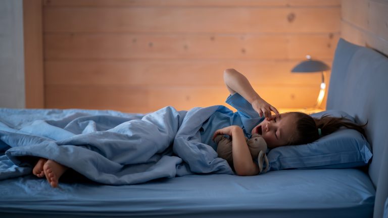 Geschenkideen zum Schulanfang, 8 clevere Produkte für einen smarten Start Ein Kind liegt im Bett bei schummrigem Licht und reibt sich verschlafen die Augen, um wach zu werden.