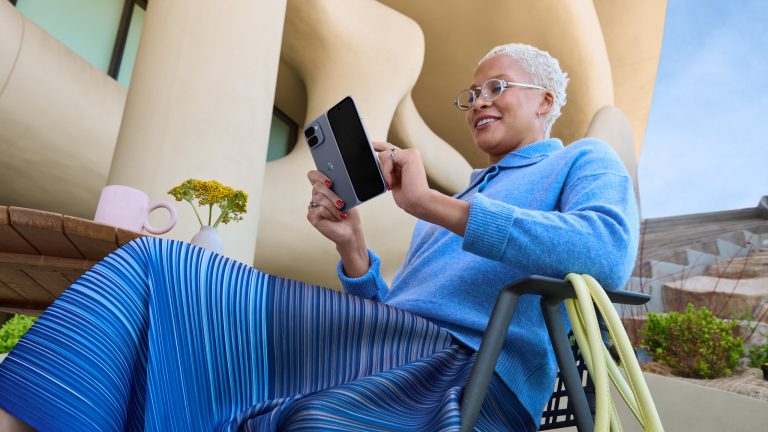 Eine Person sitzt vor einem futuristischen Gebäude auf einem Stuhl an einem Tisch und tippt auf ihrem aufgeklappten Google Pixel 10 Pro Fold.