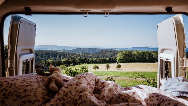 Camping Apps, Must-Haves für den Urlaub mit Camper oder Wohnmobil Blick aus dem Bett im geöffneten Heck eines Campervans auf Felder und dahinterliegenden Wald.