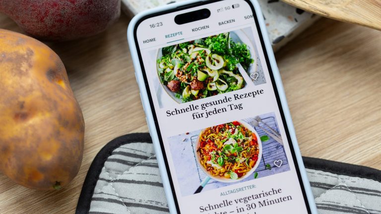Camping Apps, Must-Haves für den Urlaub mit Camper oder Wohnmobil Ein Smartphone liegt neben Kartoffeln auf einer Holzoberfläche und einem Ofenhandschuh. Darauf ist die На главную der Lecker-App mit schnellen Rezepten zu sehen.