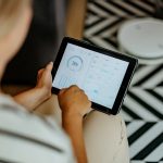 Aufnahme aus der Vogelperspektive einer Person, die ein Tablet in den Händen hält auf dessen Display eine App zur Smarthome-Steuerung geöffnet ist.