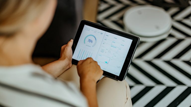 Aufnahme aus der Vogelperspektive einer Person, die ein Tablet in den Händen hält auf dessen Display eine App zur Smarthome-Steuerung geöffnet ist.