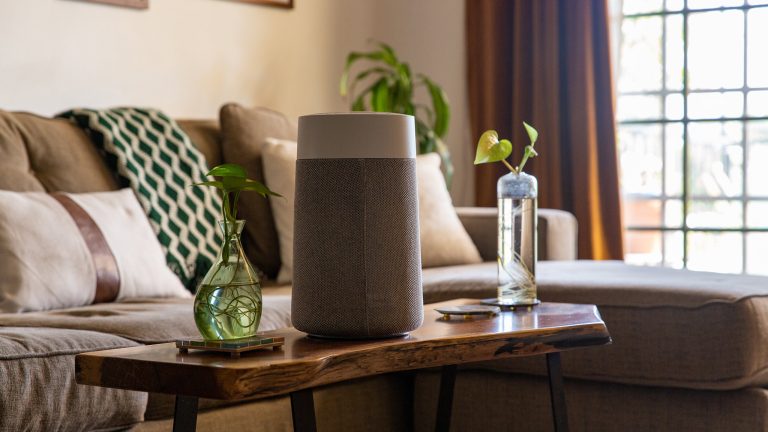 Ein Luftreiniger steht auf einem Couchtisch aus Holz. Daneben steht eine grüne Vase, im Hintergrund ist ein Sofa mit Zierkissen in Erdtönen zu sehen.