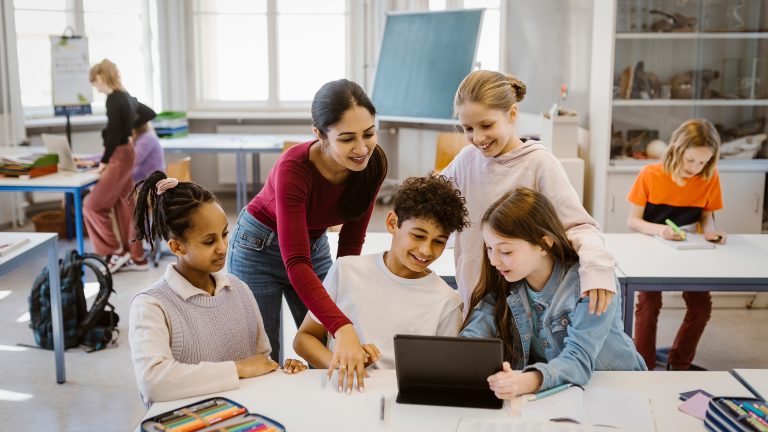 Schul-Apps, Stundenplan, Wörterbuch und Co. Eine Gruppe von Schulkindern sammelt sich um ein Tablet, das auf einem Schultisch abgestellt ist.