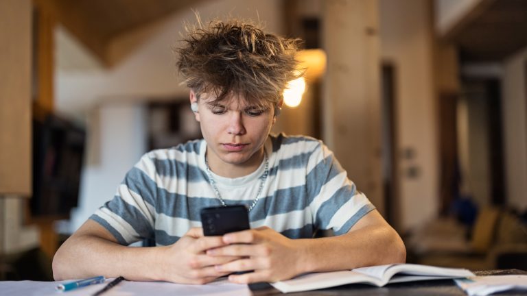 Schul-Apps, Stundenplan, Wörterbuch und Co. Ein Schulkind sitzt mit dem Handy in der Hand an einem Schreibtisch, auf dem Schulbücher liegen.