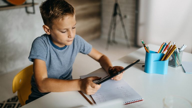 Schul-Apps, Stundenplan, Wörterbuch und Co. Ein Kind sitzt mit einem Tablet in der Hand an einem Schreibtisch, auf dem ein Block liegt.