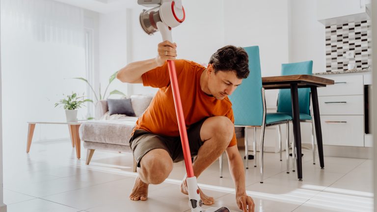 Dyson Staubsauger Alternativen, Diese Modelle überzeugen im Alltag Eine Person sitzt in der Hocke. Mit der linken Hand wischt sie mit einem Tuch den Boden, in der rechten Hand hält sie ein kabelloses Kombi-Gerät.