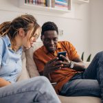 Zwei Personen sitzen nebeneinander auf einer Couch und schauen gemeinsam in ein Smartphone, das eine Person in den Händen hält.