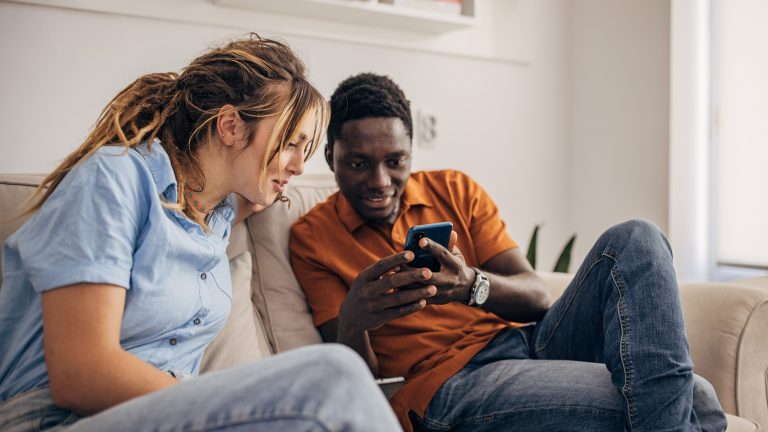 Zwei Personen sitzen nebeneinander auf einer Couch und schauen gemeinsam in ein Smartphone, das eine Person in den Händen hält.