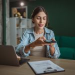 Eine Person sitzt an einem Schreibtisch. Vor ihr steht ein geöffneter Laptop, ein Klemmbrett mit Dokumenten liegt daneben. Über diese hält die Person ihr Smartphone.