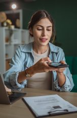Mit dem Smartphone scannen, So digitalisierst du Papierdokumente und Fotos ganz einfach Eine Person sitzt an einem Schreibtisch. Vor ihr steht ein geöffneter Laptop, ein Klemmbrett mit Dokumenten liegt daneben. Über diese hält die Person ihr Smartphone.