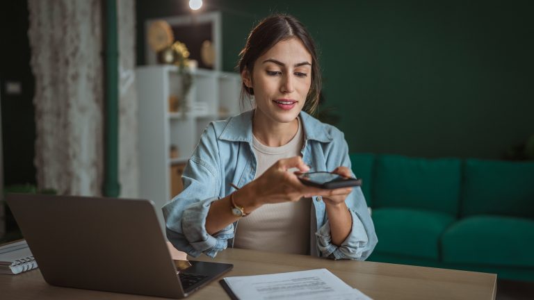 Eine Person sitzt an einem Schreibtisch. Vor ihr steht ein geöffneter Laptop, ein Klemmbrett mit Dokumenten liegt daneben. Über diese hält die Person ihr Smartphone.