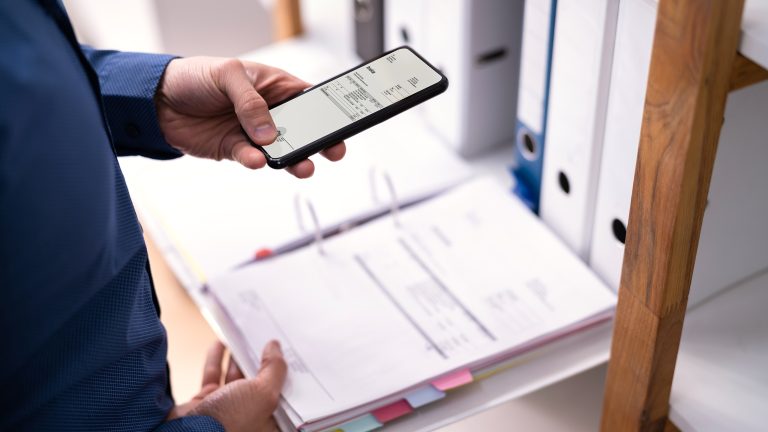 Nahaufnahme der Hand einer Person, die ein Smartphone über einen geöffneten Ordner mit Dokumenten, hält.