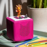 Eine pinke Toniebox steht auf einer grünen Ablage. Im Vordergrund liegt buntes Papier und Buntstifte.