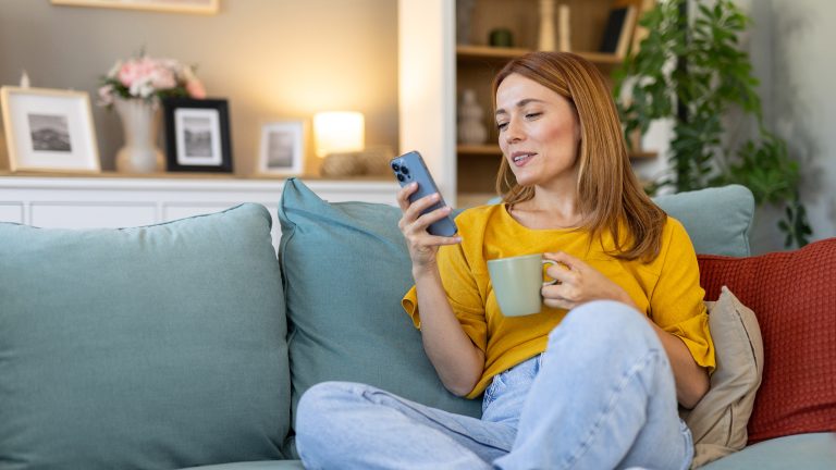 Eine Person sitzt im Schneidersitz auf einem Sofa und hält eine Tasse in der einen und ihr Smartphone, auf das sie blickt, in der anderen Hand.