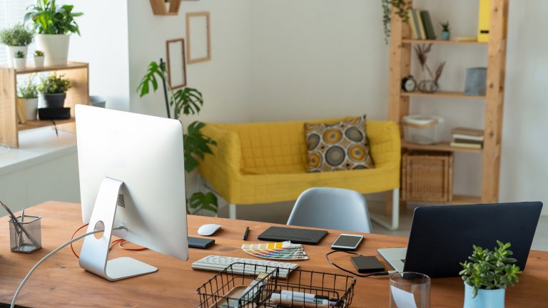 Ein Blick auf ein leeres Arbeitszimmer mit heller, hölzerner Einrichtung, vielen Pflanzen und einem zentralen Schreibtisch mit Laptop und Bildschirm.