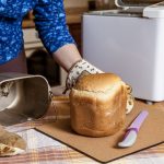 Eine Person hat gerade mit behandschuhten Händen ein frisch gebackenes Weißbrot aus dem Backbehälter eines Brotbackautomaten geholt. Das liegt auf einem Brett mit einem Messer neben dem Automaten.
