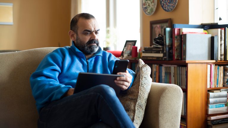 WhatsApp & iPad, So klappt es mit der Kombination Eine Person sitzt mit überschlagenen Beinen auf einem Sofa. In der linken Hand hält sie ein Smartphone, ein Tablet liegt auf ihrem Schoß.